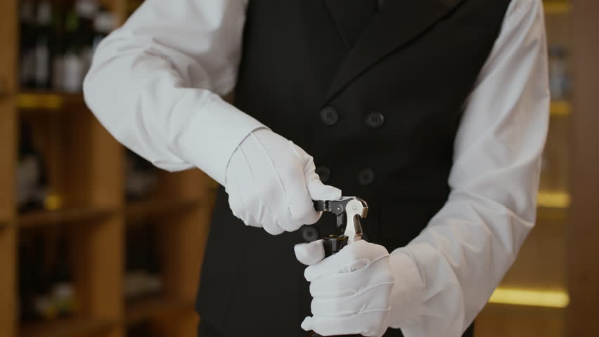 Midsection shot of hands of unrecognizable waiter or sommelier wearing elegant suit vest and white textile gloves pulling cork out of bottle using corkscrew working, in fine dining restaurant
