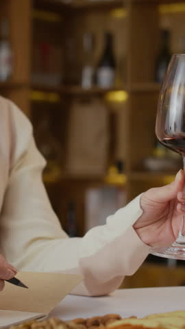 Vertical of focused young Caucasian female sommelier filling out degustation card while tasting red wine at work