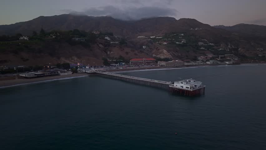 Sunset view of Malibu Pier, California, showing surfers near the lagoon outlet with Pacific Coast Highway and the coastal mountains behind. Soft dusk colors reflect across the water and shoreline.
