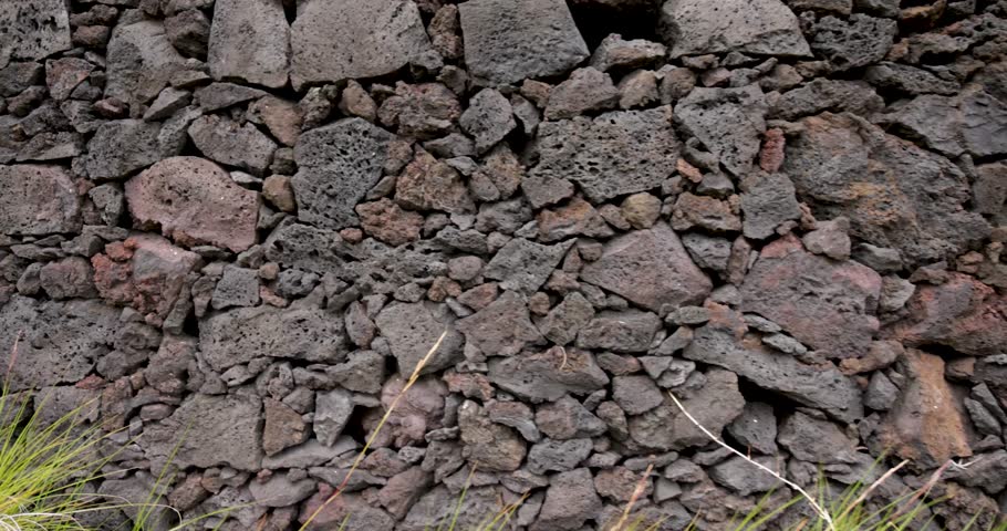 A wall made of volcanic rocks and a grassy area in front of it. The wall is made of large rocks and the grass is short. Trucking shot.