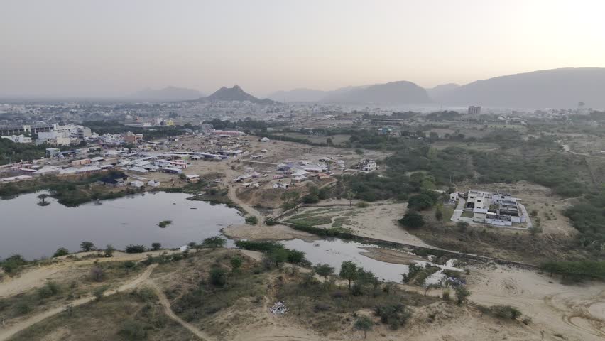 Aerial view of a pond in the desert area of Pushkar, Rajasthan during the cattle fair, showing camels, tents, and the Aravalli mountains in the background at sunrise.