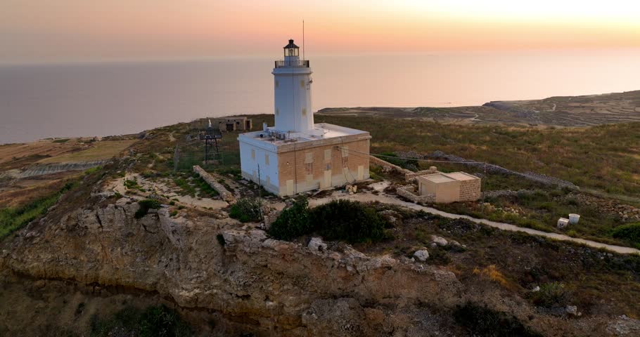 Aerial view of the white Ta' Gurdan Lighthouse standing tall on the rugged Gozo coastline against the backdrop of a serene sunset, Ghasr, Gozo, Malta.