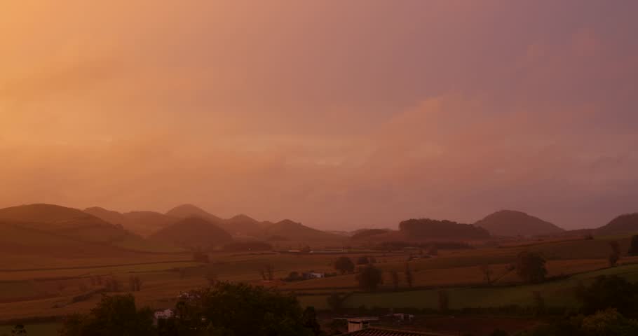 A beautiful sunset over a small town with houses and a hill at Capelas, Azores. The sky is saturated orange and the sun is setting. Panning left shot.