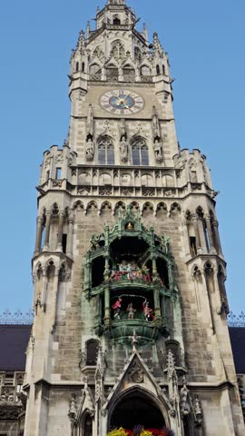architectural facade under bright blue sky, sunlit stonework and floral displays adorn historic clock building, vibrant flags and sculpted statues accentuate grand gothic clock tower scene. Munich