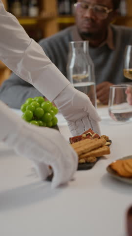 Vertical of unrecognizable waiter in elegant white gloves offering delicious appetizers for customers sitting at table in luxurious restaurant, holding platter with figs and breadsticks