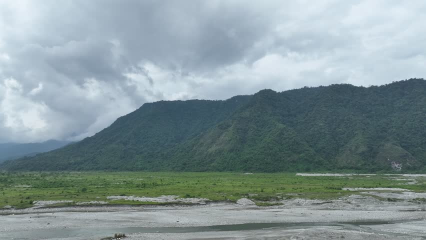 Wide aerial view of a large riverbed with flowing water, surrounded by mountains under a cloudy sky. Dramatic nature landscape with rugged terrain.