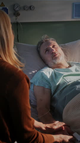 Woman with long blonde hair sits beside elderly man lying in hospital bed. Daughter holds father hand sharing quiet moment in softly lit hospital room