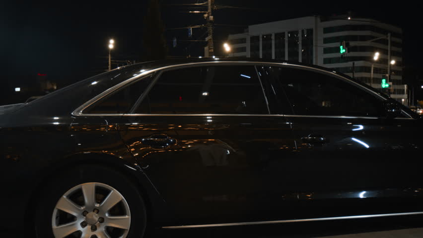 Middle aged woman in white suit getting into back seat of luxurious car while male personal chauffeur assisting VIP passenger with opening door and loading shopping bags into vehicle in night city