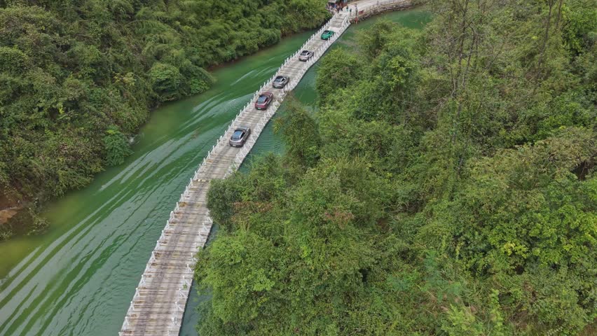 Aerial drone shot of the Shiziguan floating bridge in Hubei, China, showing cars crossing over green river water surrounded by dense forest and mountains.