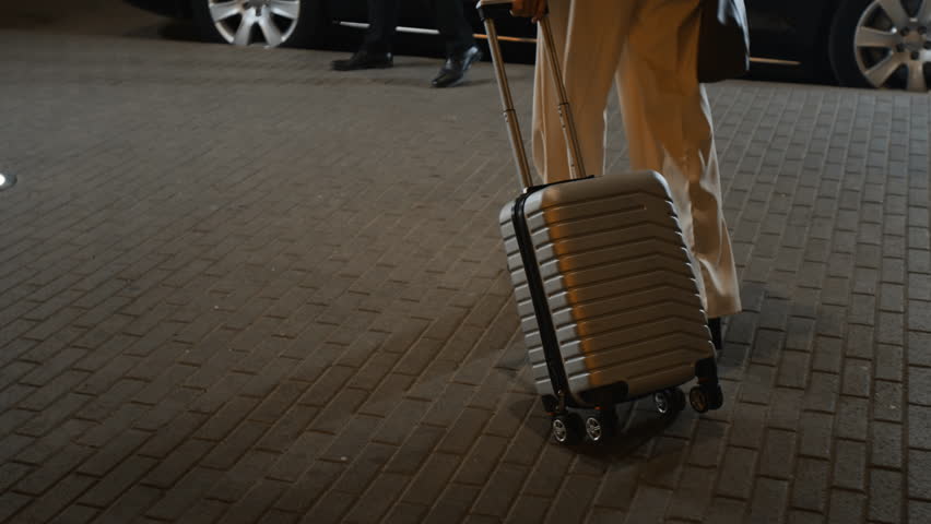Cropped shot of successful businesswoman carrying suitcase to premium class car while male personal driver assisting with opening door and loading baggage, business client leaving hotel in evening