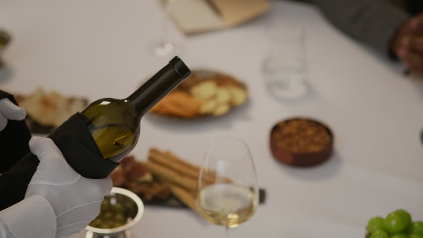 High angle cropped slowmo shot of hands of unrecognizable waiter in white textile gloves serving white wine at dinner in luxurious restaurant