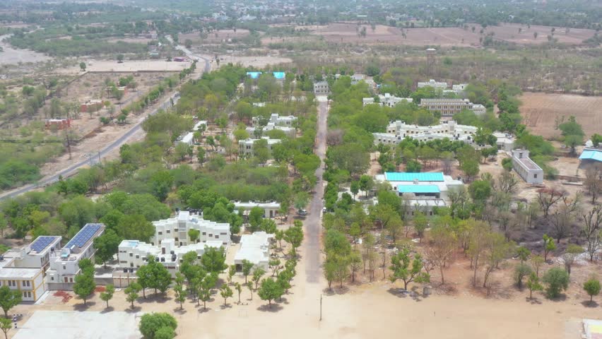 Drone footage showing a large campus with white buildings, solar panels, blue rooftops, a swimming pool, and a central road layout in a semi-arid landscape.