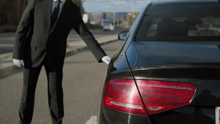 Adult male personal driver in chauffeur cap opening car door for businesswoman leaving automobile in city street, providing premium service and safe transportation for important client