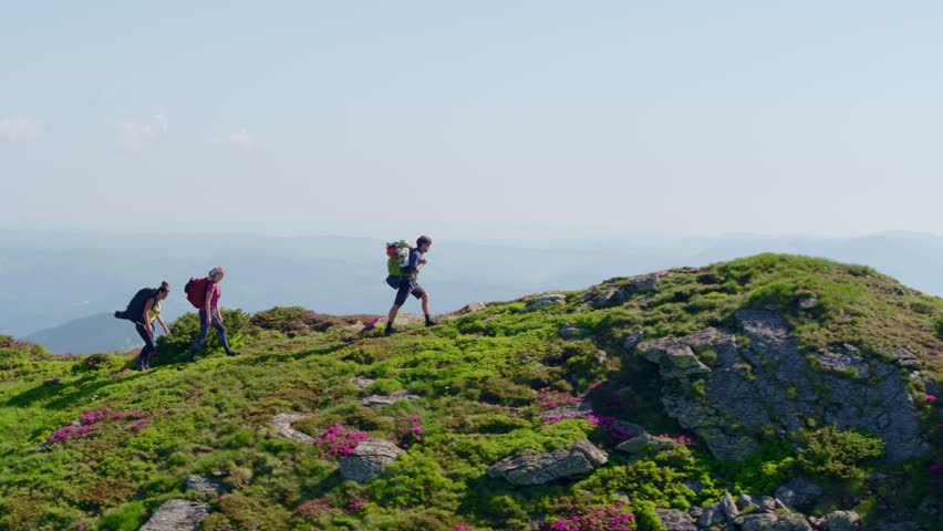 Aerial shot of three backpackers reaching a mountain summit viewpoint. Conveys adventure, achievement, freedom, and extreme sports. Filmed on a clear, sunny day.