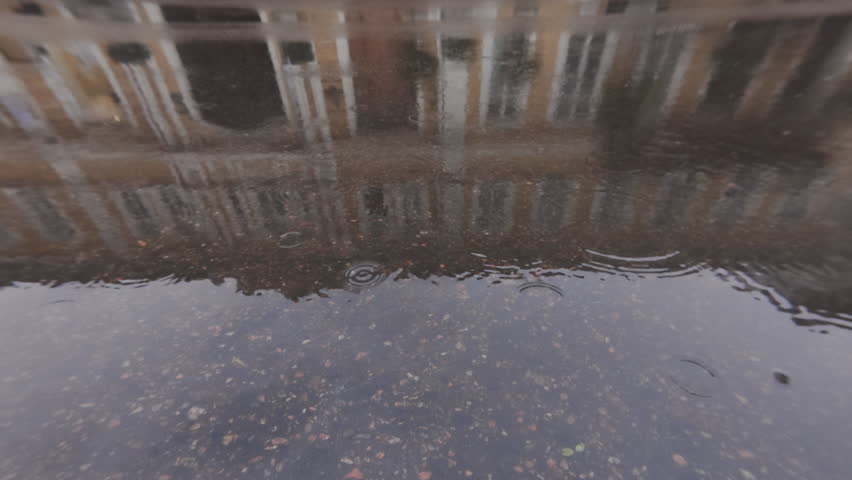 Car drives along an asphalt road during rain, automobile and houses of old town are mirrored on surface of water, drops falling on water blur these reflections. Mirroring in puddles during rain