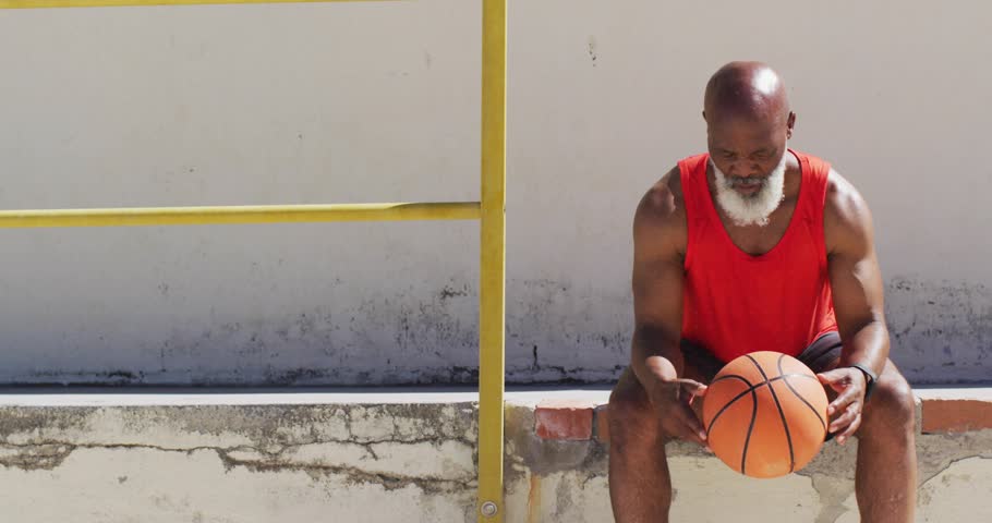 Bearded man sitting on steps with yellow rail holding ball, sun glinting causing animated arc promo. Basketball, athletic, outdoor, concrete, handrail, sunlit, urban
