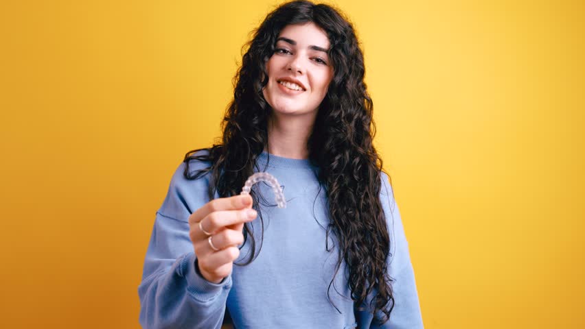 Happy young woman with long curly hair holding and pointing at a transparent removable aligner for dental correction on a yellow background