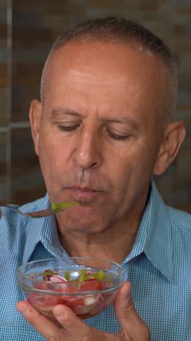 Portrait of a 60-year-old Man Eating a Mixed Fresh Salad at his Home. A Middle-aged man in a blue shirt sitting at the dining table, enjoying a mixed vegetable salad dressed with olive oil.