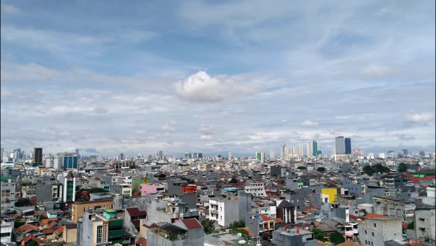 Jakarta cityscape panorama with a clear blue sky and scattered white clouds. Capturing a dense urban area with a mix of high-rise skyscrapers and residential buildings.