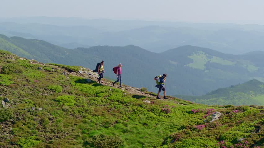 Aerial shot of three trekking along a scenic, grassy mountain ridge with backpacks. Conveys active lifestyle, friendship, and outdoor exploration on a clear day.