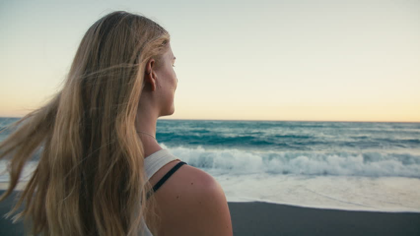 Rear medium close-up of a woman with flowing blonde hair as she gazes at the sea, her hair gently lifted by the wind at sunset.