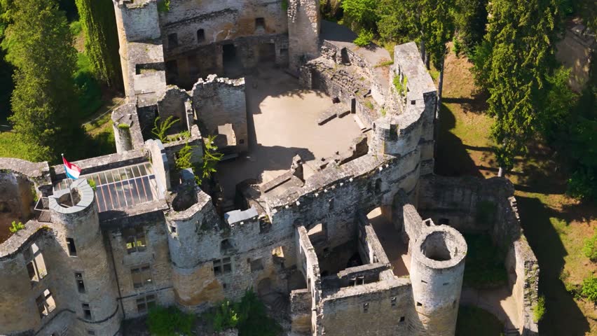 medieval castle beaufort outer walls ruins green gardens flag tall trees traffic road outside parked cars luxembourg descending closeup circling top drone shot