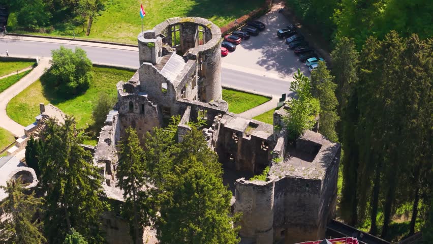 medieval castle beaufort outer walls ruins green gardens flag tall trees parked cars visitors arriving luxembourg closeup circling top drone shot les châteaux de