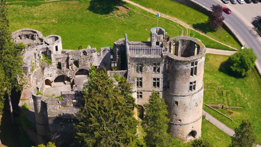 medieval castle beaufort outer walls ruins green gardens flag tall trees people walking inside luxembourg closeup circling top drone shot les châteaux de