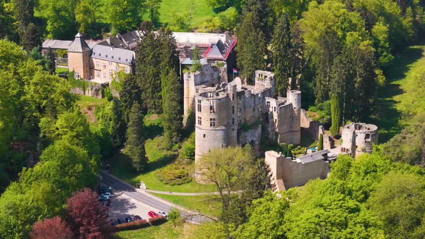 medieval castle beaufort outer walls ruins green gardens flag tall trees parked cars people walking luxembourg to left around right top drone panning les châteaux de