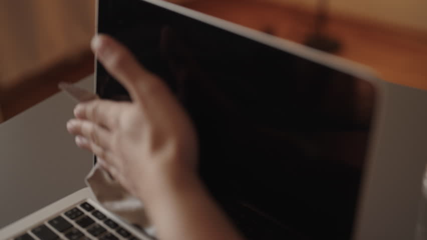 Unrecognizable person wiping dusty laptop screen with microfiber cloth after spraying cleaning solution, promoting hygiene and proper electronic device maintenance for optimal performance, closeup.