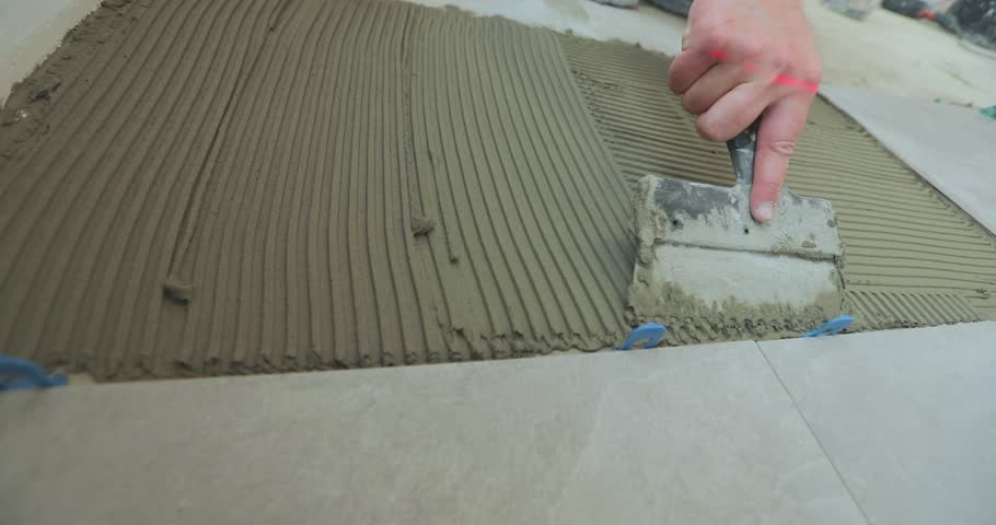 Laying Ceramic Tiles. Troweling adhesive onto a concrete floor in preparation. Applying thin set tile mortar on a tile. Install Ceramic Tile Flooring, close-up. 