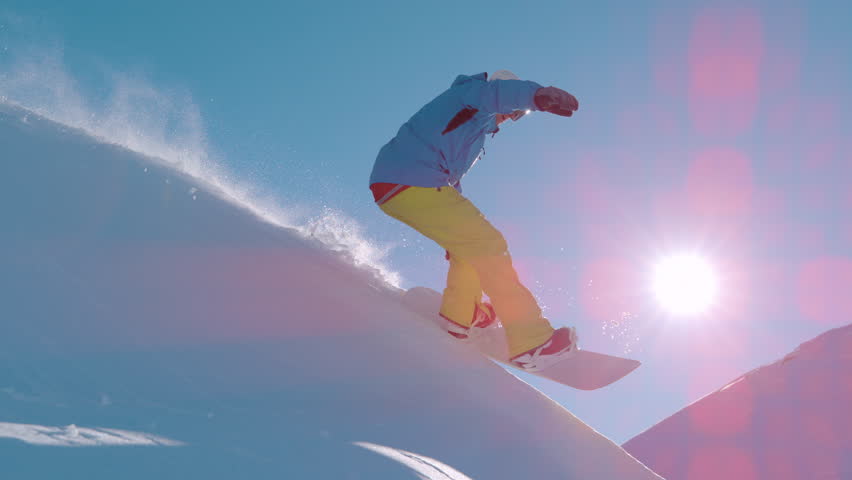 SLOW MOTION, LENS FLARE: Snowboarder carves down a steep powder slope, sunbeams fill the frame. Fresh snow sprays through air as skilled freerider playfully rides down the snowy mountains in the Alps.