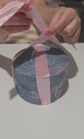 Close-up vertical shot of hands tying a pink ribbon bow around a round gift box; elegant holiday present wrapping, Christmas, Valentine