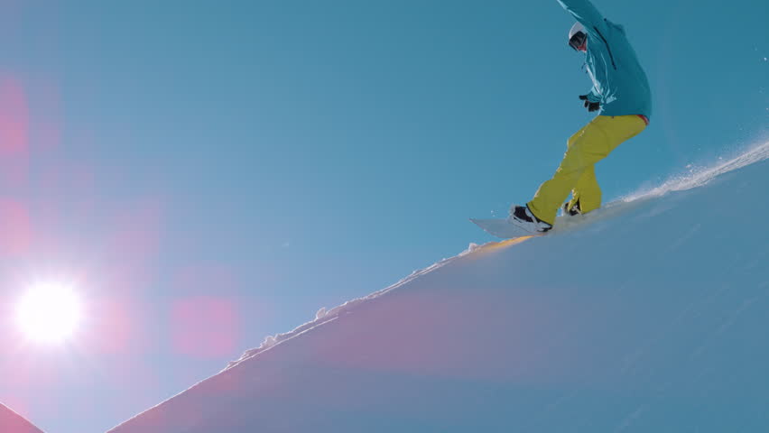 SLOW MOTION, LENS FLARE: Snowboarder leans into a powerful slash turn high above a sunlit peak, kicking a sparkling plume of powder as he crashes. Playful backcountry freeriding in the European Alps.