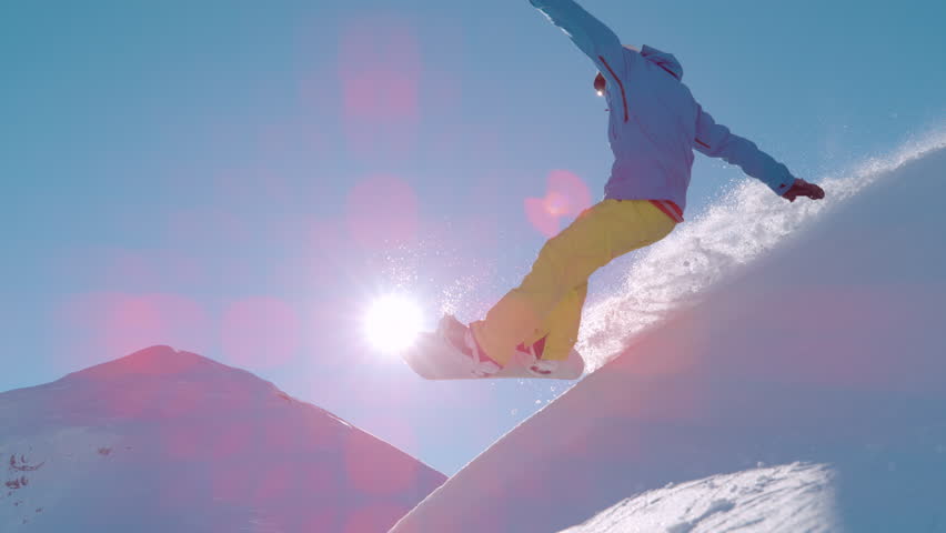 SLOW MOTION, LENS FLARE: Snowboarder leans into a powerful slash turn high above a sunlit peak, kicking a sparkling plume of powder as he crashes. Playful backcountry freeriding in the European Alps.