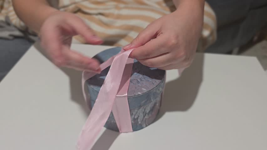 Horizontal close-up video of hands carefully tying a pink ribbon bow around a stylish round gift box; elegant wrapping for Christmas, Valentine