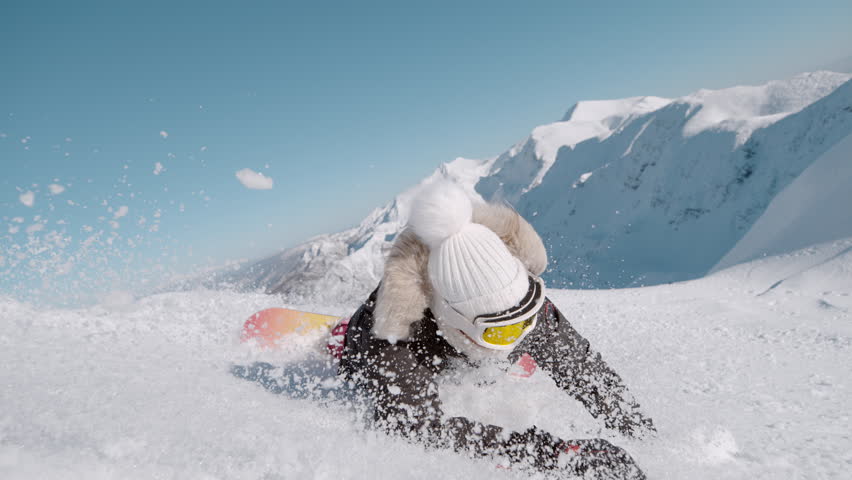 SLOW MOTION, CLOSE UP: Female snowboarder tumbles into soft snow on a sunny alpine slope as she tries to learn to carve on a snowboard. Snow flies toward the camera while she crashes with a smile.