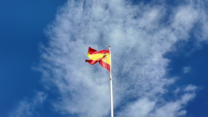 Spanish flag waving in the wind against a bright blue Mediterranean sky.