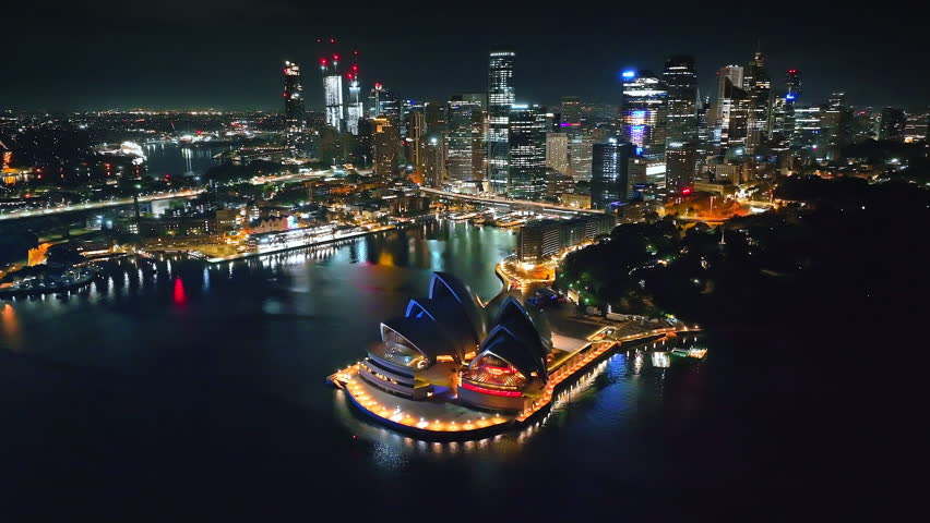Australia, Sydney Opera House glowing brightly at night, city harbor surrounded by illuminated skyline with skyscrapers reflecting lights on water. Travel destination. Aerial drone flight footage