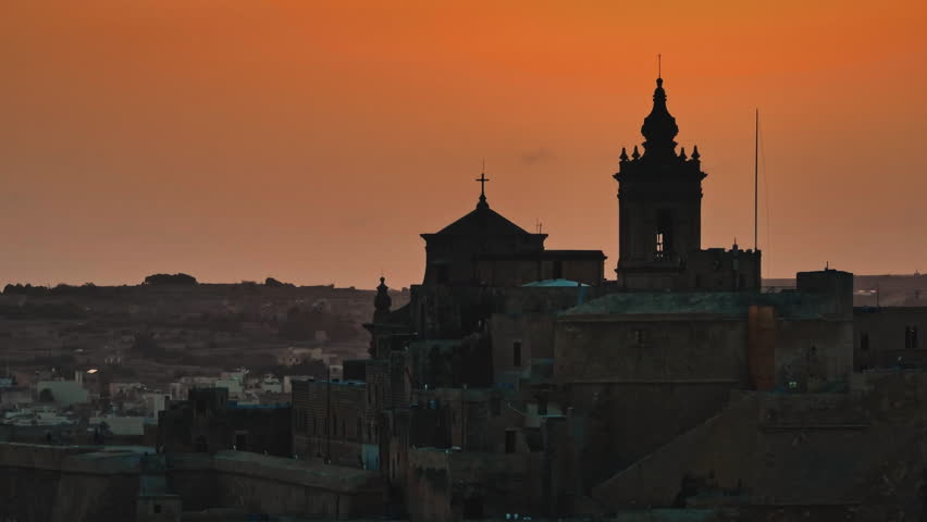 Malta, Comino island: Illuminating the silhouette of the Cathedral of the Assumption in the Cittadella, an orange sunset casts a warm glow over Victoria, Gozo. Drone flight footage