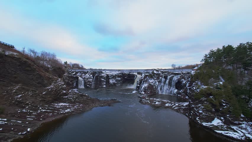 Cinematic FPV aerial view of a winter waterfall flowing between snowy rocks. Cold tones, mist, frozen textures, natural landscape and dramatic winter atmosphere.