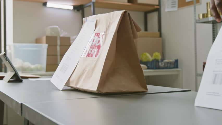 Close-up of hands of anonymous African American employee placing packed bag with stapled invoice, containing grocery order, on counter at dark store or online supermarket, ready for delivery or pickup