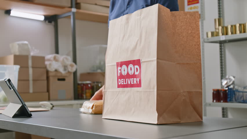 Medium shot of young Caucasian female employee placing bag of dry cereal in paper package, then walking to rack, picking up apples, while collecting goods to fulfill order at dark store