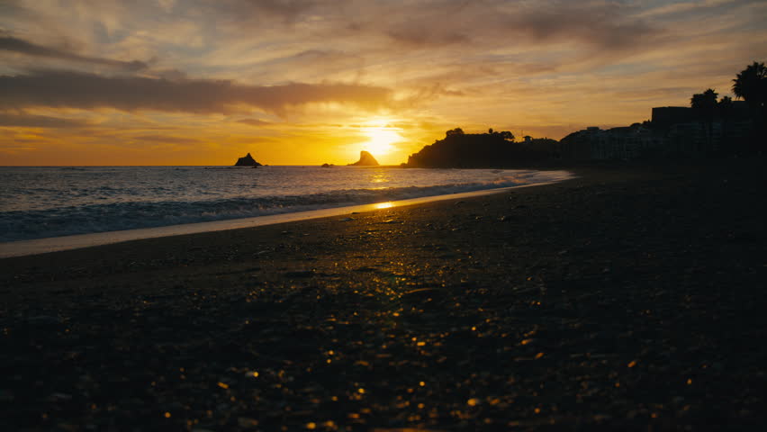 Dramatic sunset over a dark coastal beach in Spain. Golden light reflects off wet sand with moody, cinematic atmosphere.