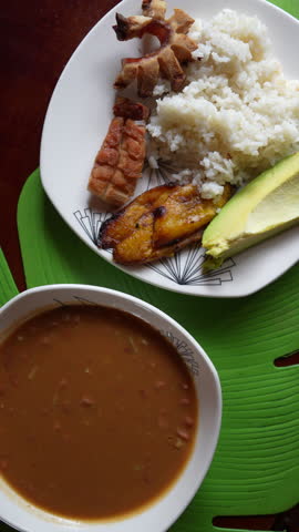 Vertical video showcasing a vibrant close-up shot of traditional Colombian bandeja paisa with savory beans, crispy pork, rice, plantain, and avocado. Perfect for mobile social media.