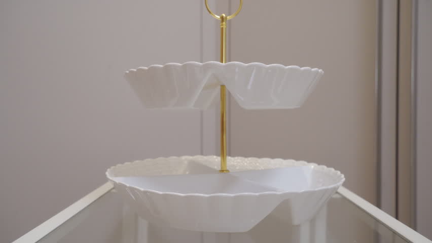 White glossy ceramic and gold metal tiered serving dish is set down on a glass surface. A light, delicate clink rings out.