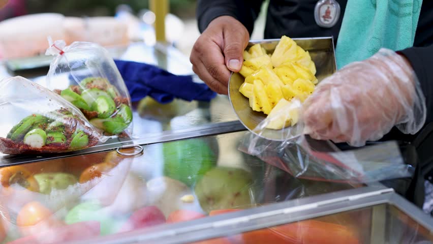 Gloved vendor places pineapple chunks into plastic bag at colorful outdoor fruit market cart