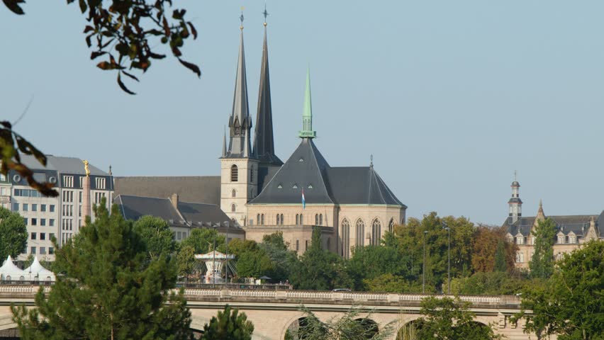 Static wide shot of historic cathedral, city buildings, bridge, and autumn trees under clear daylight