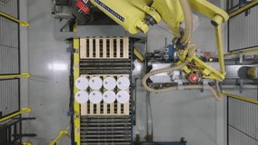 Top-down aerial shot of a yellow robotic arm performing palletizing by stacking white pails and buckets onto a wooden pallet on an automated production line. - Powered by Shutterstock - Get 15% off with code: PIKWIZARD15