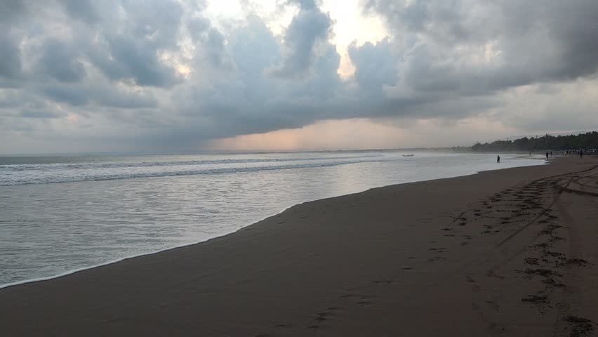 Majestic beach sunset under dramatic, cloudy sky. Waves gently roll onto the wide, sandy shore with footsteps and distant figures. Peaceful, moody coastal beauty